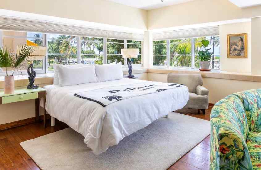 Quarto de um hotel em Miami Beach em frente à praia, com cama de casal, tapete, poltrona, sofá, luminárias, plantas decorativas e janelas grandes ao redor