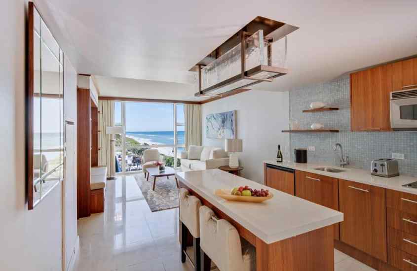 Sala com cozinha conjugada de hotel em Miami Beach com sofá, poltrona, mesas, cadeiras, quadros decorativos e janela grande acortinada