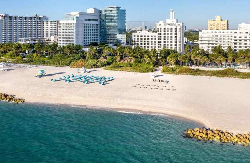 Em um dia de sol, vista aérea de hotel em Miami Beach com praia privativa, guarda-sóis coloridos e árvores atrás