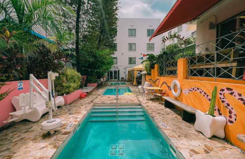 Em um dia de sol, área de lazer de hotel em Miami Beach com piscinas, cadeiras, bancos, árvores, plantas e pinturas coloridas ao redor