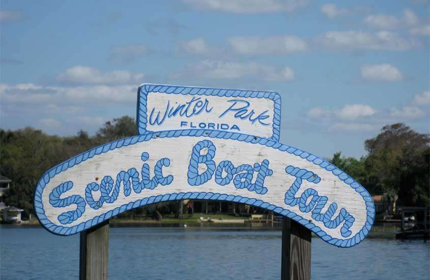Em um dia de sol com nuvens, placa do Scenic Boat Tour um dos possíveis passeios em orlando com árvores ao redor e lago atrás