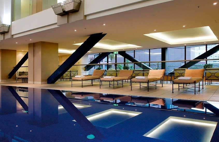 Área de lazer de hotel com piscina, espreguiçadeiras, janelas grandes e toalhas