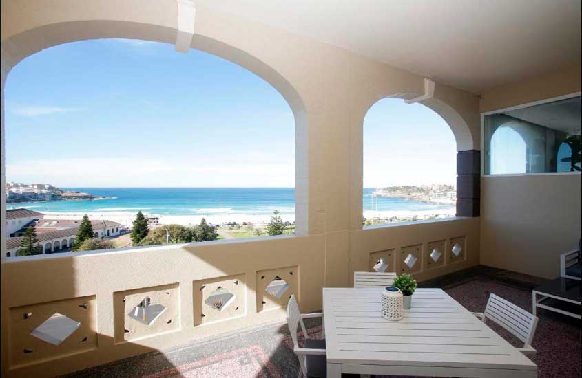 Varanda de um hotel onde ficar em Sydney com mesa, cadeiras, flores decorativas e paisagem da praia