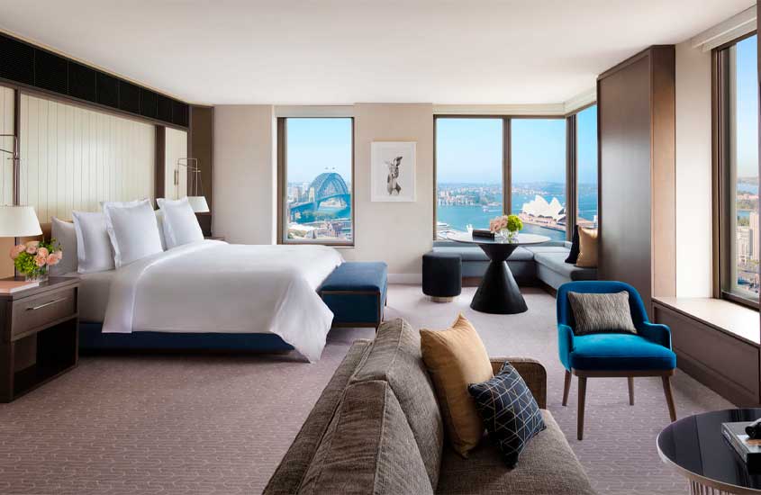 Quarto de um dos hotéis em Sydney com cama de casal, sofá, cadeiras, janelas grandes com vista da cidade, TV e mesa