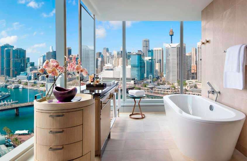 Banheiro de um dos hotéis em Sydney com janelas grandes, banheira, armários, flores decorativas, toalhas e espelho grande