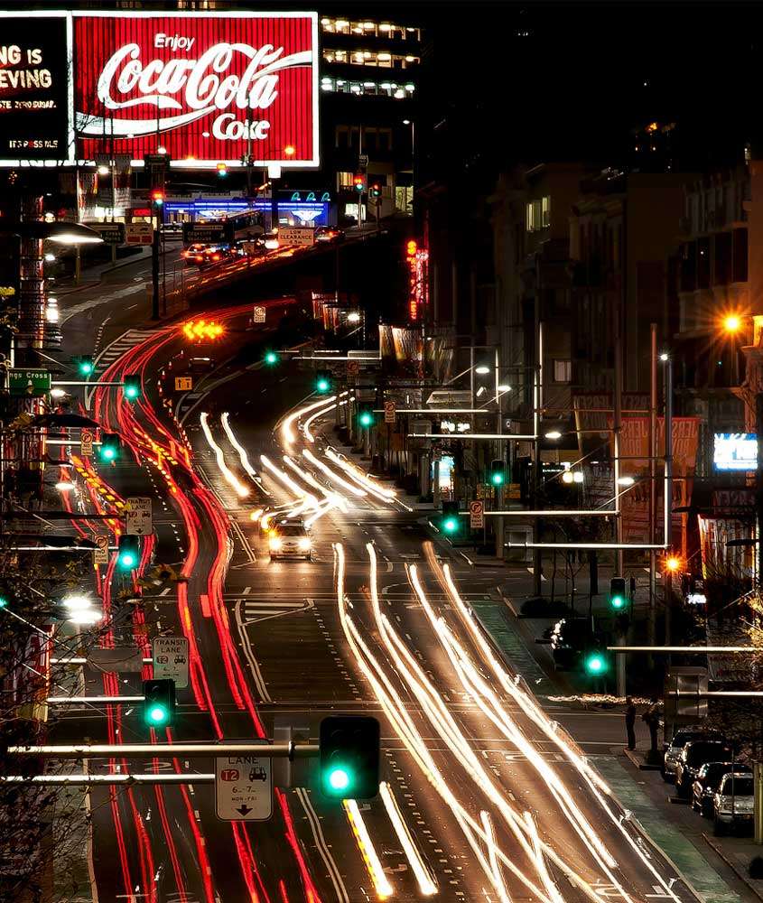 Durante a noite, rua movimentada com faróis luminosos,outdoor da coca e carros andando
