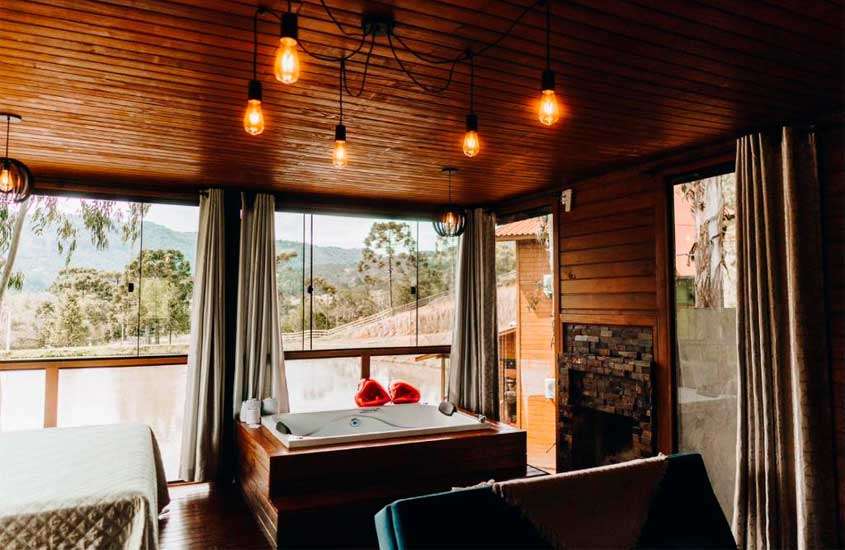 Interior de cabana na serra catarinense com cama de casal, banheira de hidromassagem, lareira e janela grande acortinada
