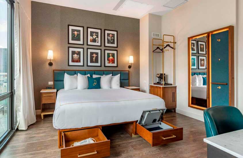 Quarto de hotel com cama de casal, gavetas, criados de madeira, quadros decorativos, espelho e mesa de trabalho