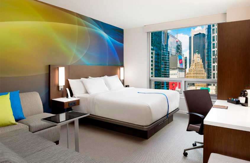 Quarto de hotel perto da Times Square com cama de casal, sofás, mesa de trabalho, parede decorada e janela grande com vista para a cidade