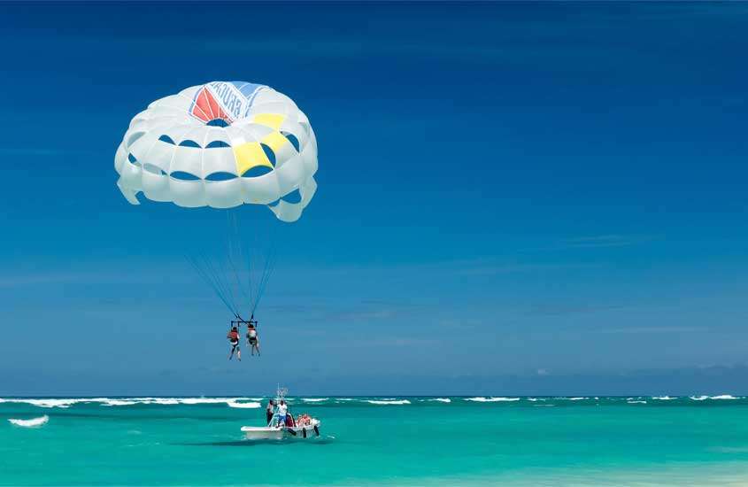 Em um dia de sol, praia com mar azul e pessoas voando de parasil, um dos pontos turísticos em punta cana