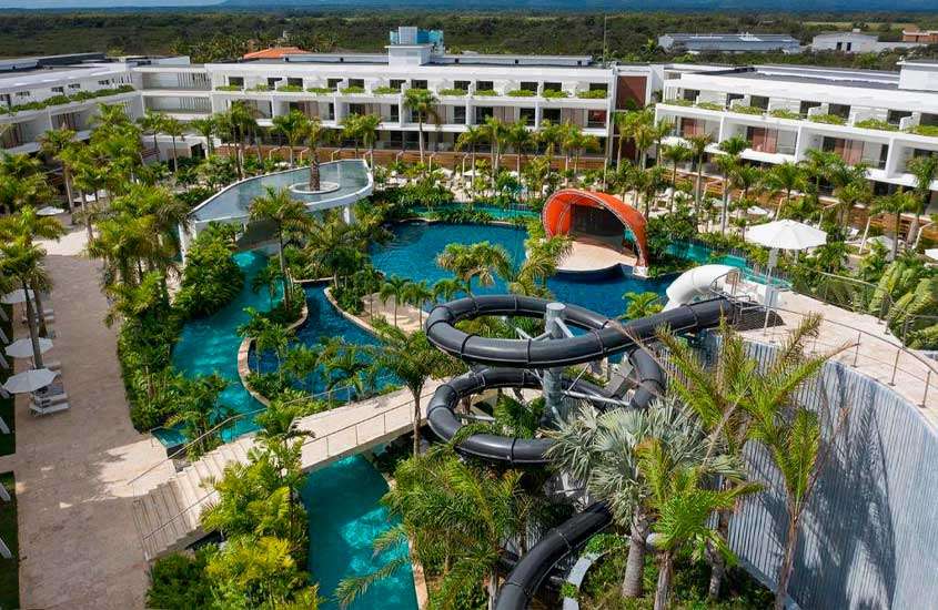Em um dia de sol, vista aérea de área de lazer de hotel com piscinas, toboágua, árvores e plantas ao redor