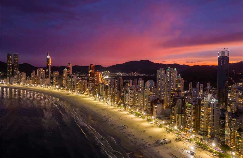 Durante o entardecer, vista aérea de Balneário Camboriú, um dos lugares para passar o réveillon com pra na frente e prédios altos iluminados atrás