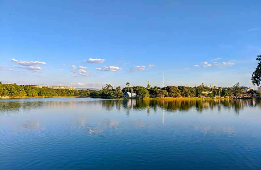 Em um dia ensolarado, paisagem da lagoa da pampulha em BH, uma das cidades para passar o réveillon, com igrejinha no meio, árvores ao redor e mineirão do lado