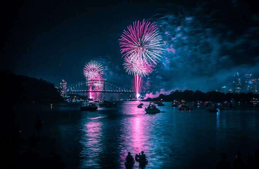 Durante a noite, queima de fogos coloridos com ponte atrás, mar e barcos em Sidney, um ótimo destino onde passar o réveillon
