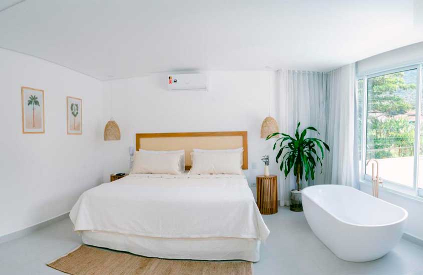 Quarto de hotel com cama de casal, tapete, banheira, planta decorativa, janela grande acortinada, ar condicionado e quadros decorativos