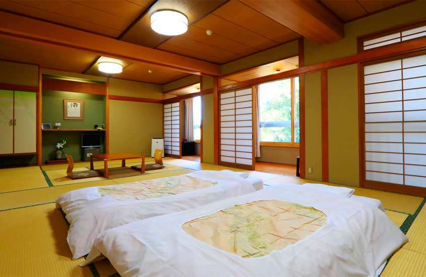 Interior de um Ryokan com camas tradicionais, mesa, cadeiras e janelas grandes