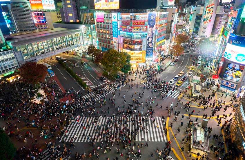 Durante a noite, pessoas no cruzamento de shibuya com árvores ao redor, painéis luminosos, carros e prédios iluminados