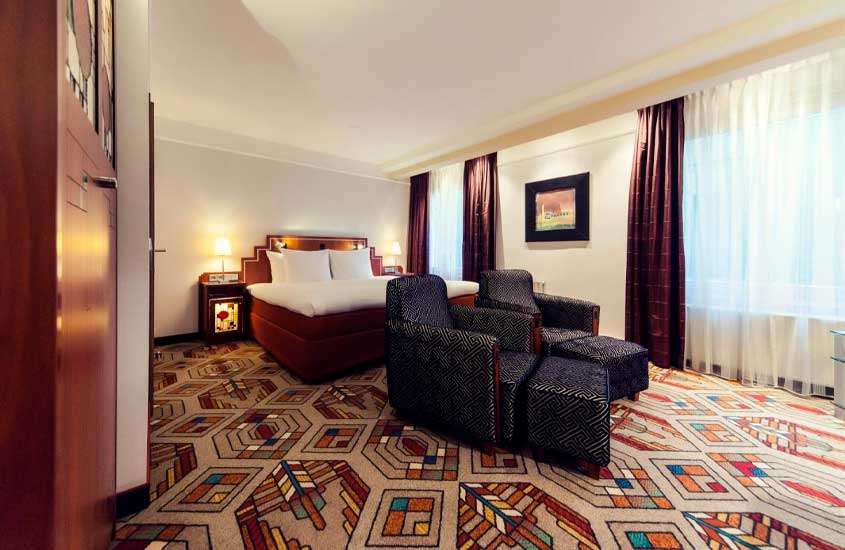 Quarto de hotel em amsterdam com poltronas, quadro decorativo, armário e janelas grandes acortinadas