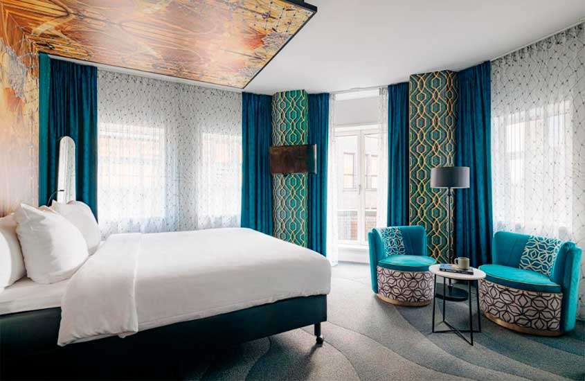 Quarto de hotel em amsterdam com cama de casal, poltronas, luminária, TV e janelas grandes acortinadas