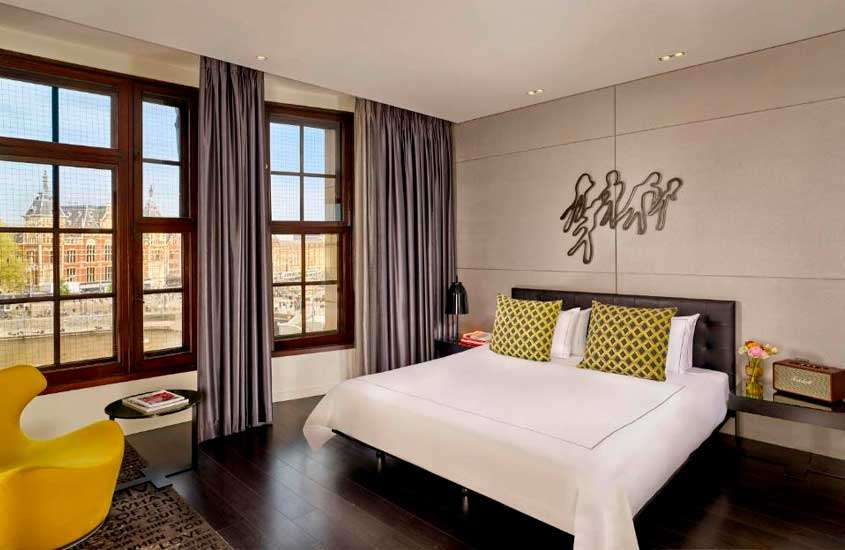 Quarto de hotel com cama de casal, poltronas, tapetes, mesa e janelas acortinadas