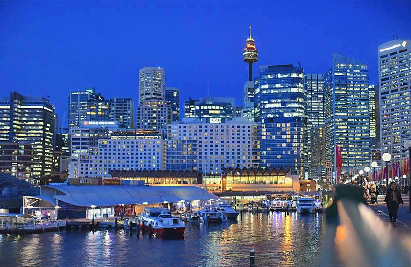 Durante o anoitece, cidade de Sydney iluminada com barcos aportados e pessoas ao redor