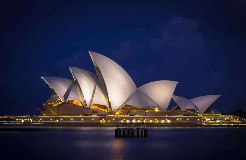 Durante a noite, visão de um dos pontos turísticos de Sydney iluminado por luzes brancas e amarelas
