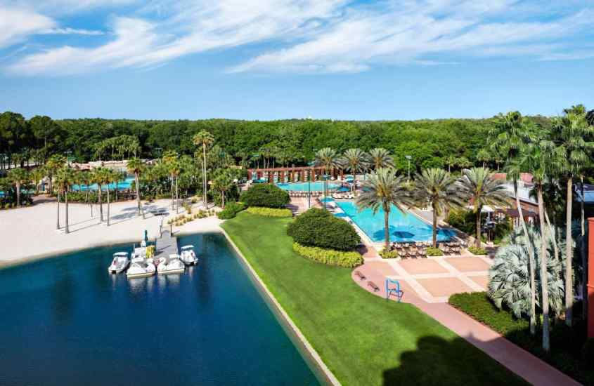 Em um dia de sol, visão aérea de um hotel para passar o Réveillon em Orlando com piscinas, lago, pedalinhos, espreguiçadeiras e vegetação ao redor