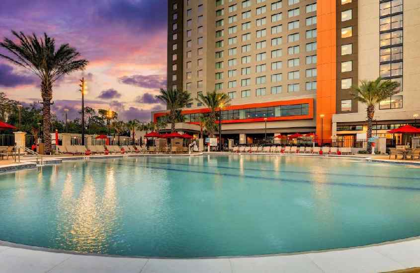 Durante o final de tarde, área de lazer de um hotel para passar o Réveillon Orlando com piscina, espreguiçadeiras, guarda-sóis e árvores ao redor
