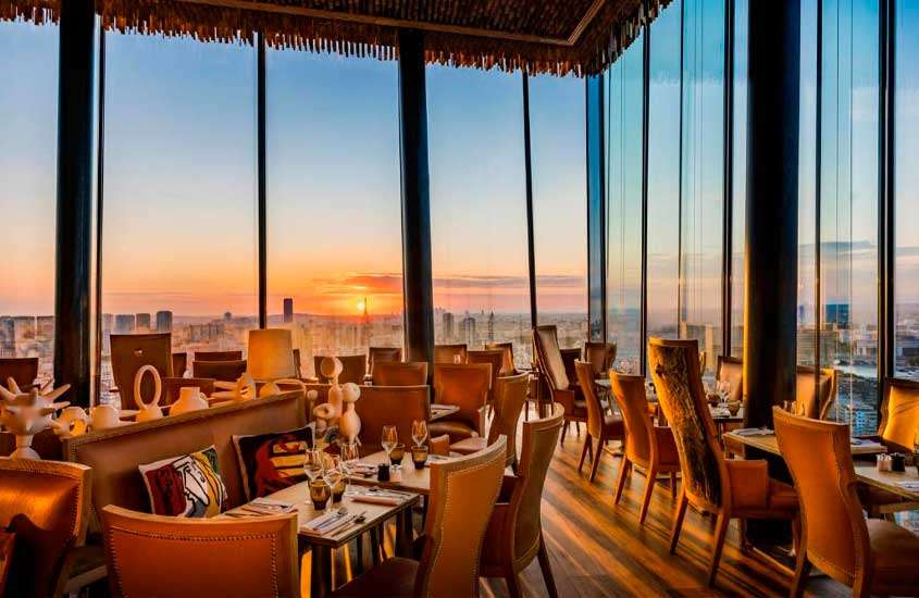 Durante o pôr do sol, restaurante com mesas, cadeiras e janelas grandes com vista da cidade