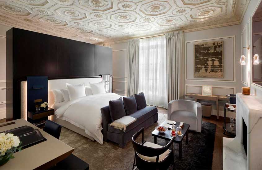 Quarto de hotel para passar ano novo em Paris com cama de casal, sofá, poltronas, tapetes, lareira, janelas grandes acortinadas, mesa de trabalho e quadro decorativo