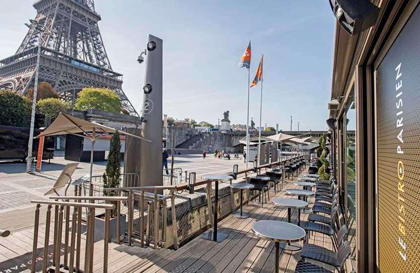 Em um dia de sol, fachada de um restaurante para passar o Réveillon em Paris com mesas, cadeiras, árvores e torre eiffel do lado