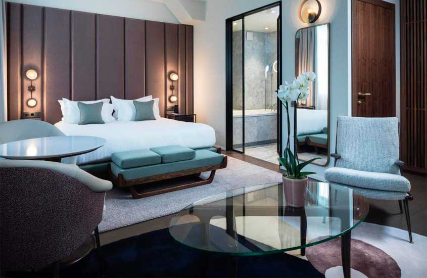 Quarto de hotel com cama de casal, poltronas, cadeiras, tapete, flores decorativas, luminárias, sofá e espelho