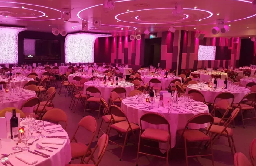 Le Duplex: Salão de eventos com luzes vibrantes em tons de rosa, mesas dispostas elegantemente com cadeiras ao redor e detalhes de decoração que criam um ambiente moderno e sofisticado