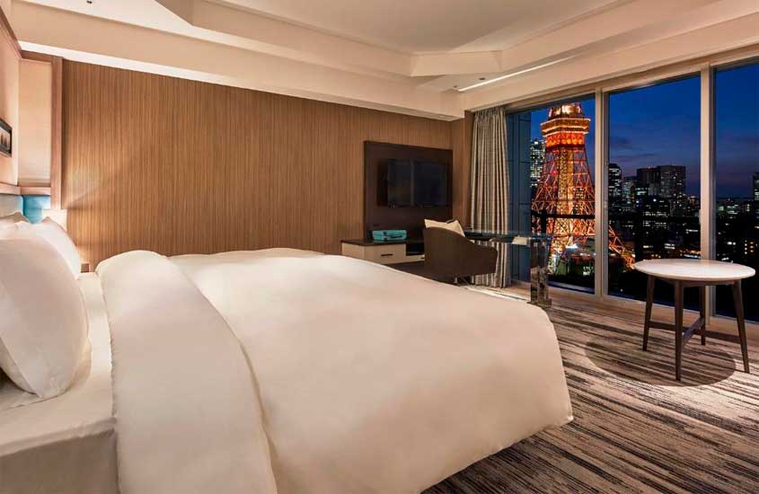 Quarto de hotel com cama de casal, TV, poltrona, mesa e janelas grandes acortinadas com vista da cidade