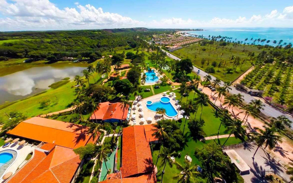 Em um dia de sol, visão aérea de um hotel fazenda em Alagoas com piscinas, lagos e árvores ao redor