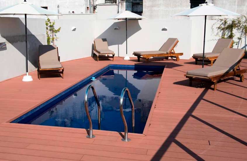 Em um dia de sol, área de lazer de um dos hotéis em Buenos Aires para brasileiros, com piscina, espreguiçadeiras de madeira, guarda-sóis e plantas decorativas