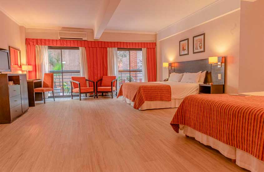 Quarto amplo de hotel com camas, mesas, cadeiras, luminárias, janelas grandes acortinadas e quadros decorativos