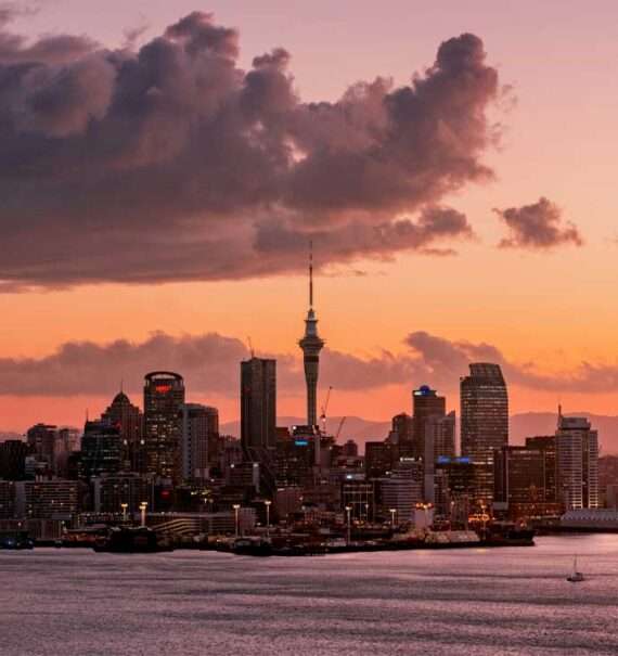 Durante o entardecer, paisagem da cidade de Auckland iluminada com navios na frente e prédios iluminados