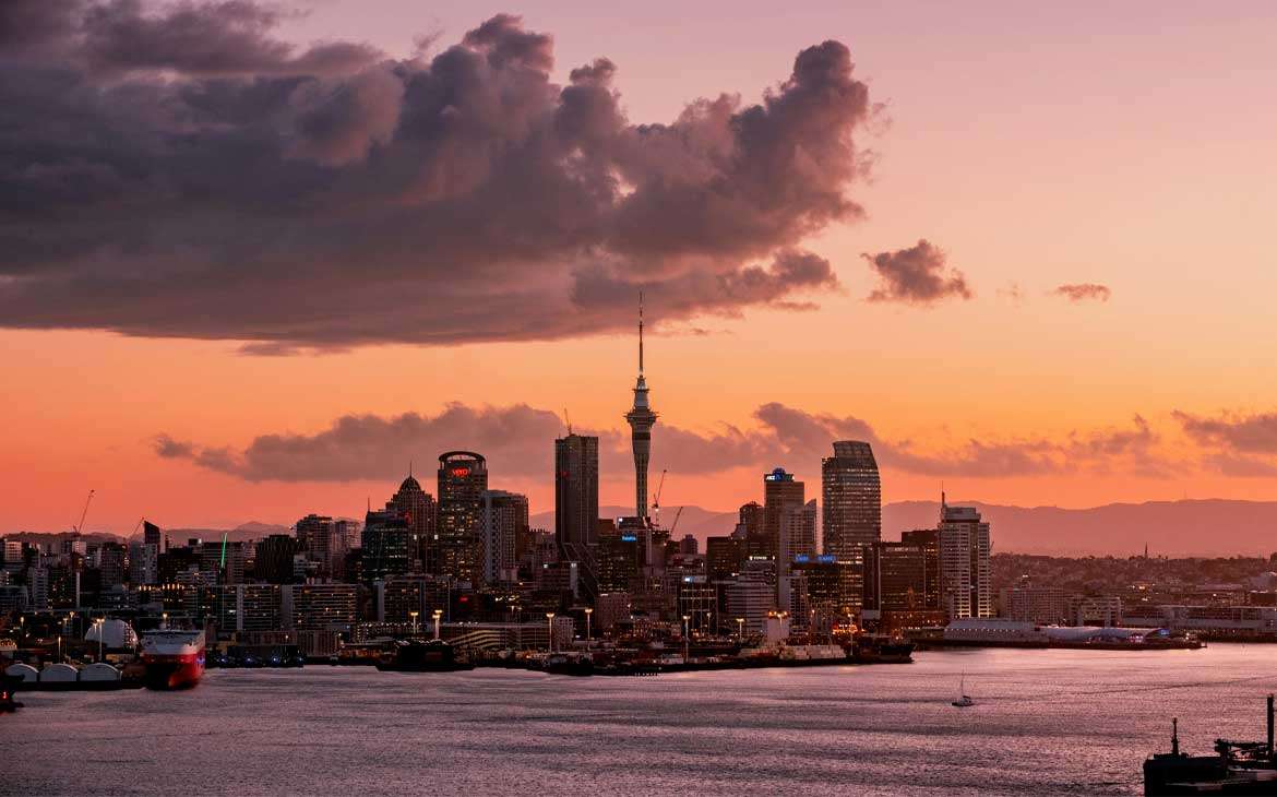 Durante o entardecer, paisagem da cidade de Auckland iluminada com navios na frente e prédios iluminados