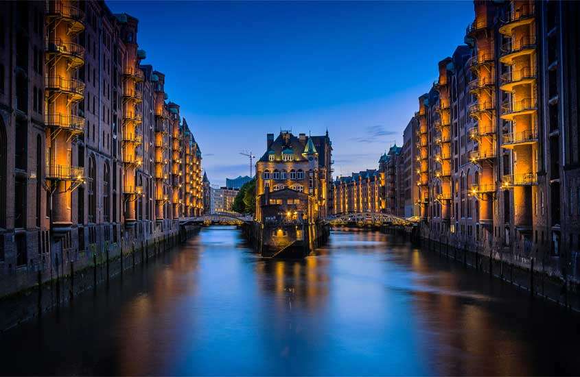 Durante o anoitecer, canais do bairro Altstadt, um dos lugares onde ficar em Hamburgo, com prédios iluminados com luzes amarelas, árvores e pontes ao redor