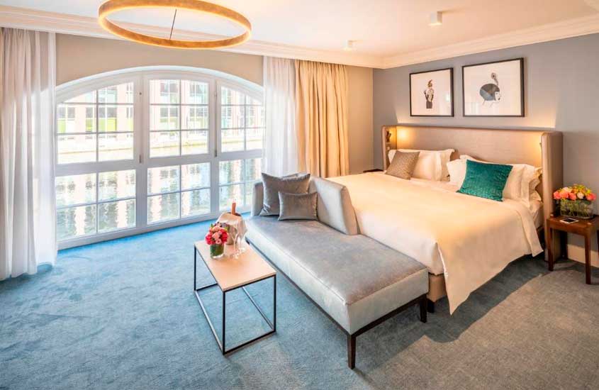 Quarto de hotel em Hamburgo com cama de casal, sofá, janela grande acortinada, lustres, flores e quadros decorativos