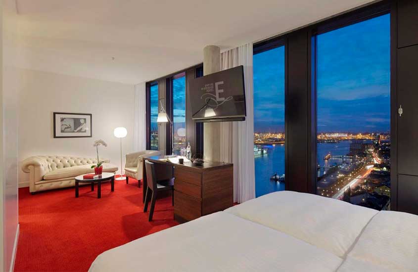 Quarto de hotel em Hamburgo, com cama de casal, TV, sofás, mesas, cadeiras, janelas grandes acortinadas, luminárias e quadro decorativo
