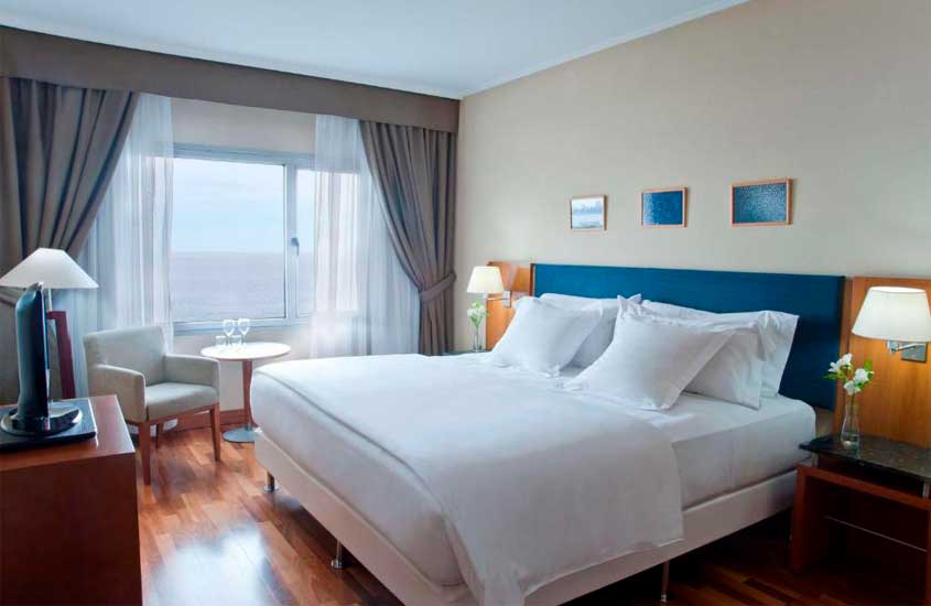 Quarto de um hotel onde passar o réveillon em Montevidéu com cama de casal, mesa, cadeiras, luminárias, quadros, janelas grandes acortinadas e plantas decorativas