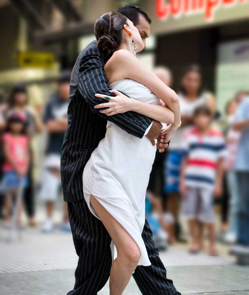 Em um dia de sol, casal em um show de tango com pessoas ao redor
