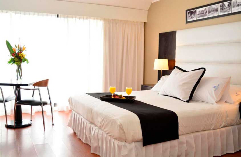 Quarto de hotel com cama de casal, mesa, cadeiras, quadros decorativos, janelas acortinadas e bandeja de café da manhã