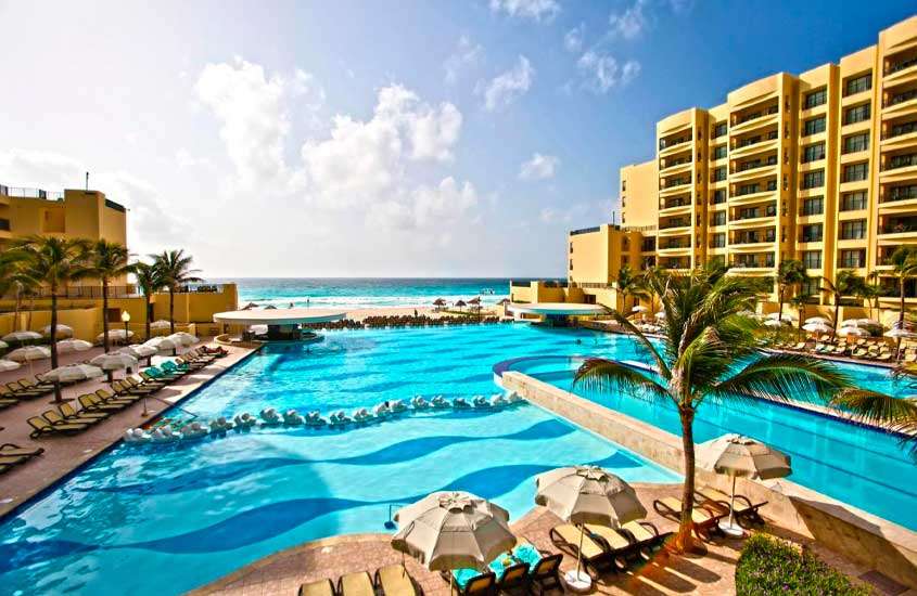 Em um dia de sol, área de lazer de hotel para passar o Réveillon em Cancún com piscina, espreguiçadeiras, guarda-sóis, árvores e praia na frente
