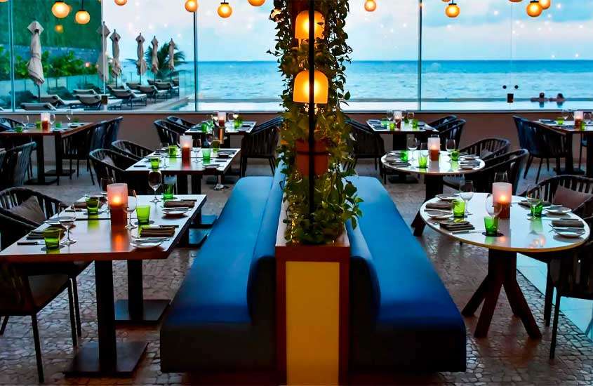 Interior de restaurante com mesas, sofás, cadeiras, flores decorativas e paisagem do mar