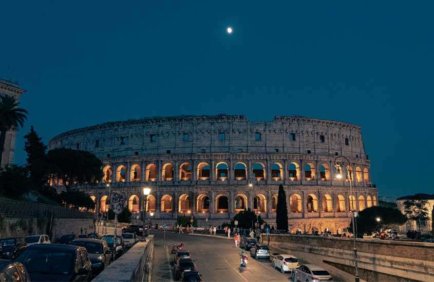 Durante a noite, Coliseu iluminado com árvores, carros e pessoas ao redor