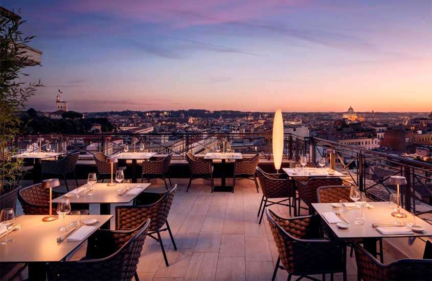 Durante o anoitecer, cobertura de um hotel para passar o Réveillon em Roma com mesas, cadeiras, plantas ao redor e paisagem da cidade