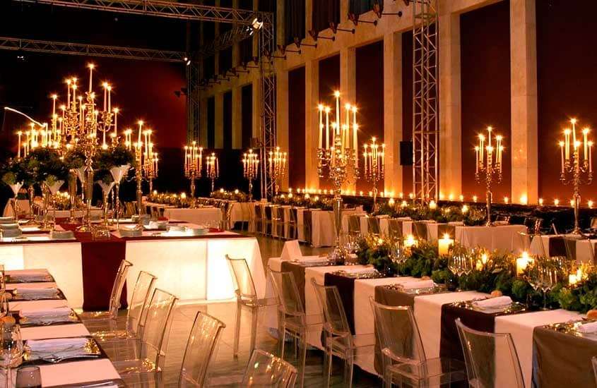 Salão de festa com candelabros, mesas, cadeiras, plantas e flores ao redor